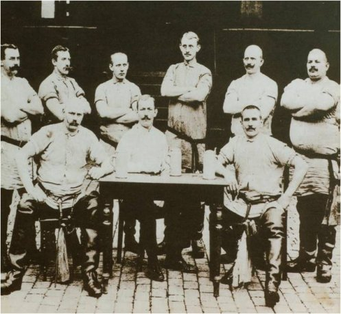 Neun Männer in Arbeitskleidung, ernst schauend, zwei sitzen an einem Tisch, schwarz-weiß Foto.