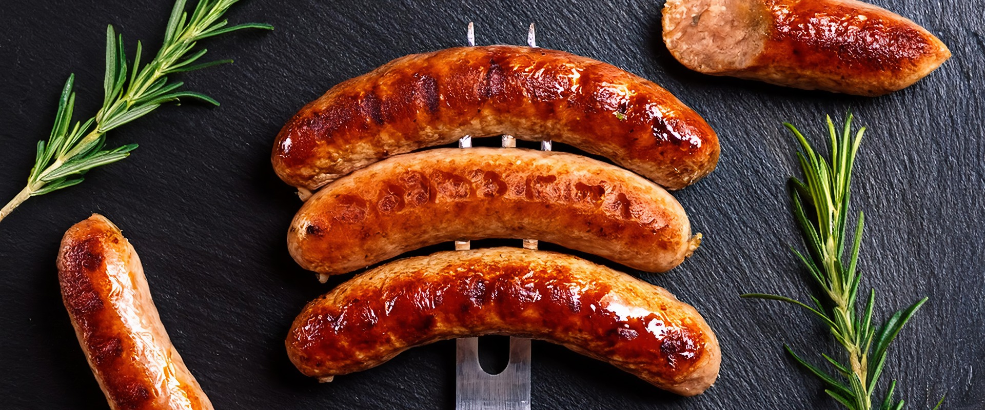 Grilled sausages on a metal meat fork with rosemary on a dark stone background. top view